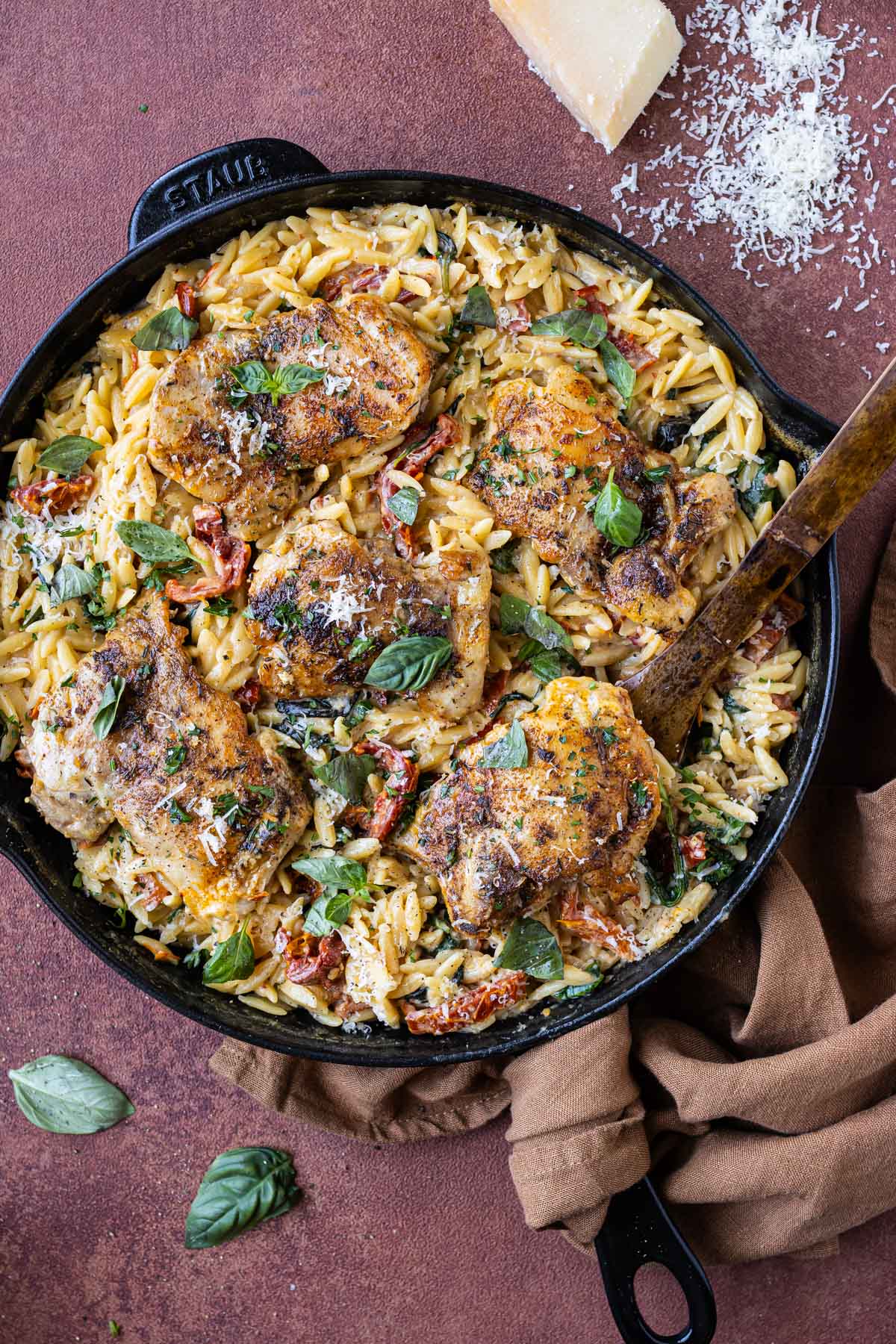 A one-pot Marry Me Chicken Orzo Pasta Bake is being served with a wooden spoon, basil, and Parmesan cheese.