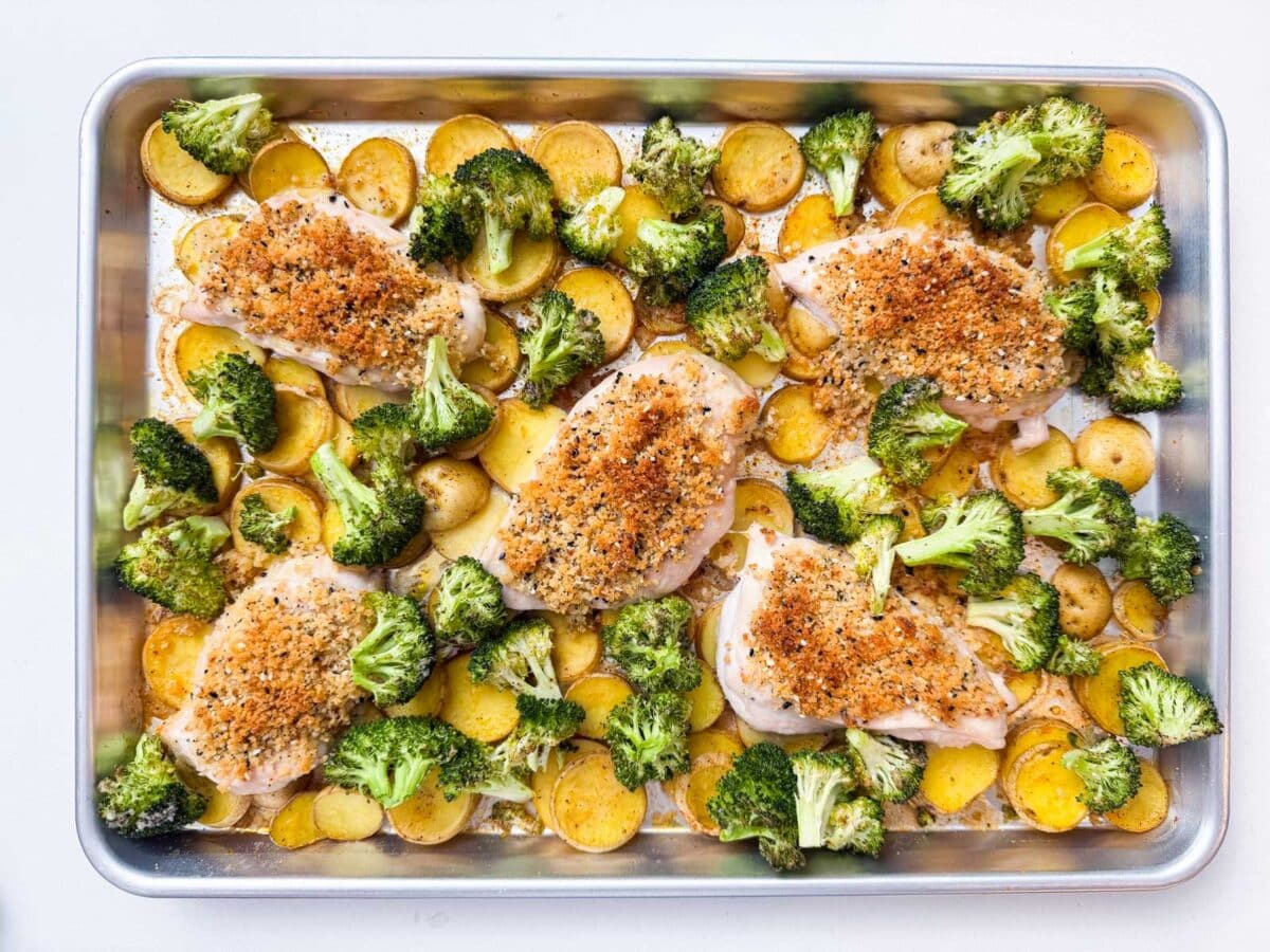 Broccoli is piled on top of sliced potatoes and chicken breasts on a sheet pan.