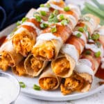 A stack of crispy baked buffalo chicken taquitos is served on a plate with Frank's Redhot and Ranch dressing drizzled on top.
