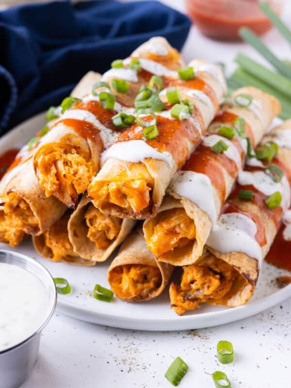 A stack of crispy baked buffalo chicken taquitos is served on a plate with Frank's Redhot and Ranch dressing drizzled on top.
