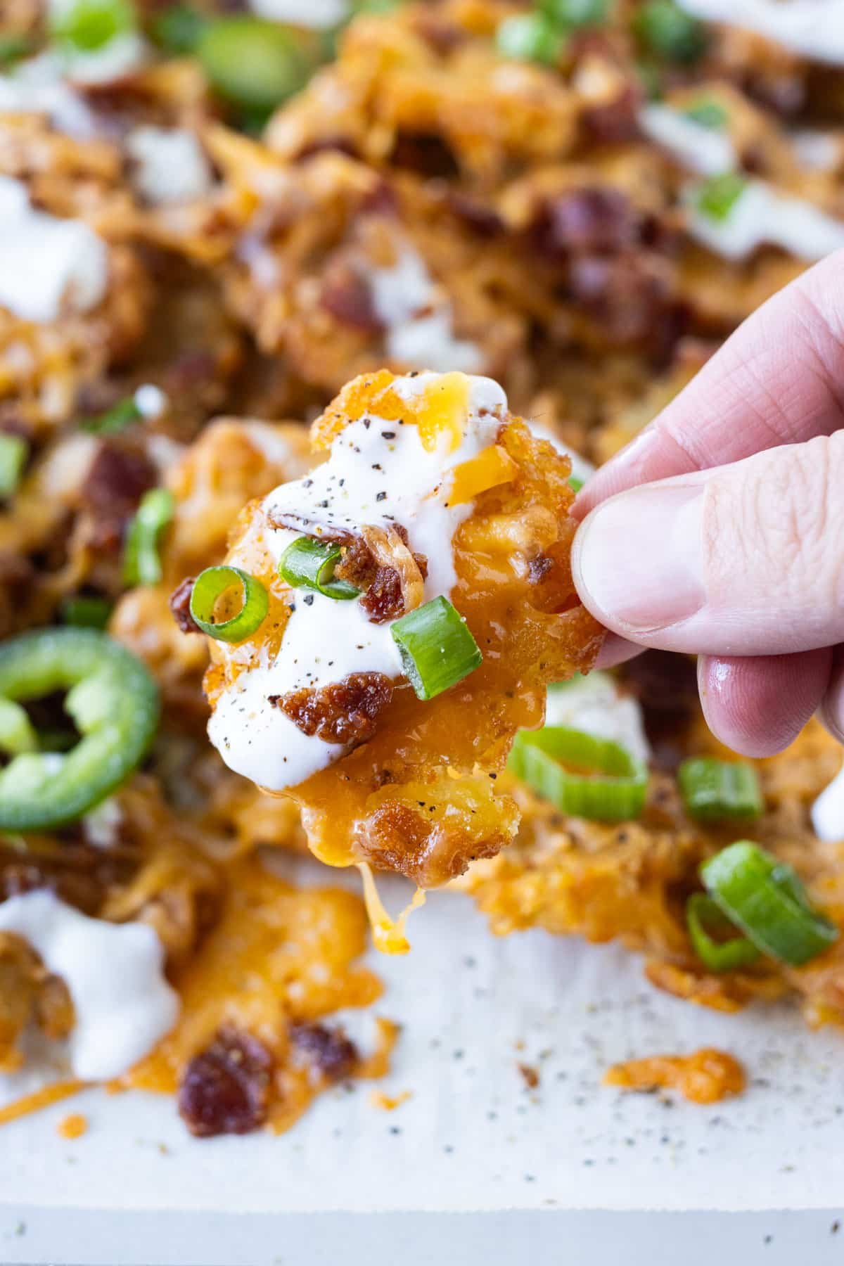 A hand is picking up a totcho, loaded smashed tater tot nacho, from a baking sheet.