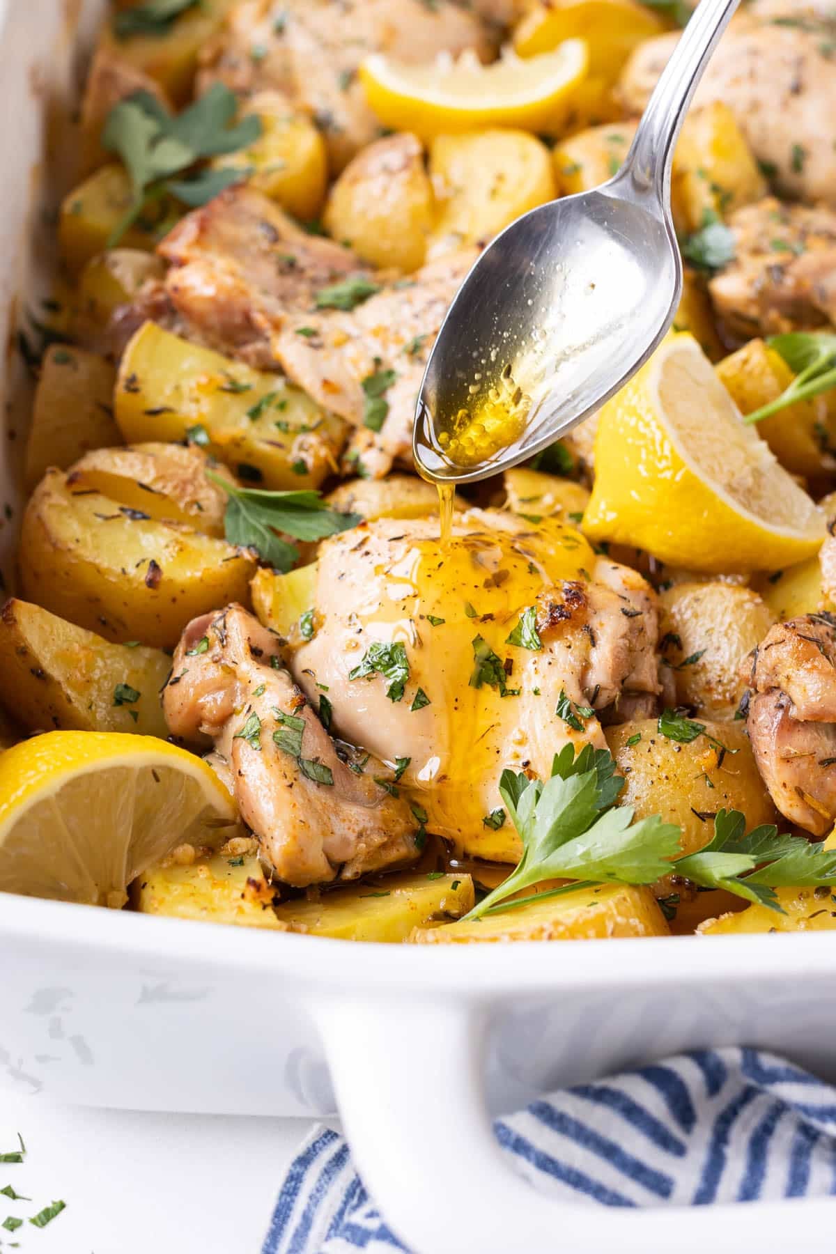A spoon is drizzling an olive oil and lemon juice pan sauce over chicken thighs and potatoes in a baking dish.