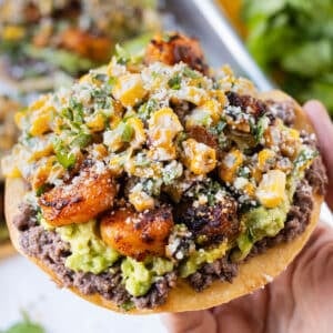 A hand is holding a Shrimp Tostada with Mexican Street Corn, avocado, and refried black beans.