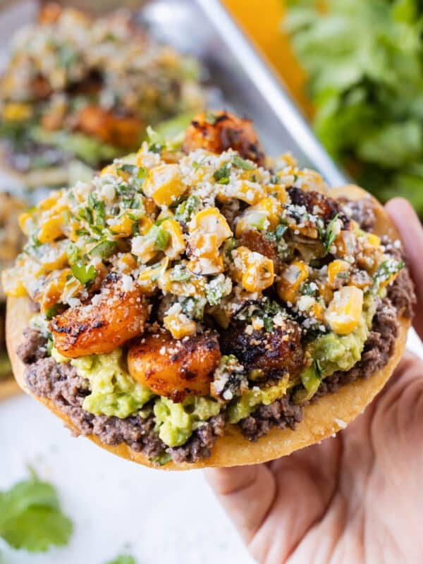 A hand is holding a Shrimp Tostada with Mexican Street Corn, avocado, and refried black beans.