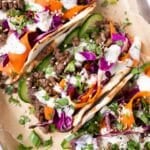 Three Korean Beef Tacos with sesame slaw are on a dinner tray next to lime wedges.