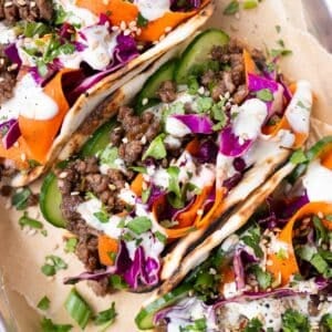 Three Korean Beef Tacos with sesame slaw are on a dinner tray next to lime wedges.