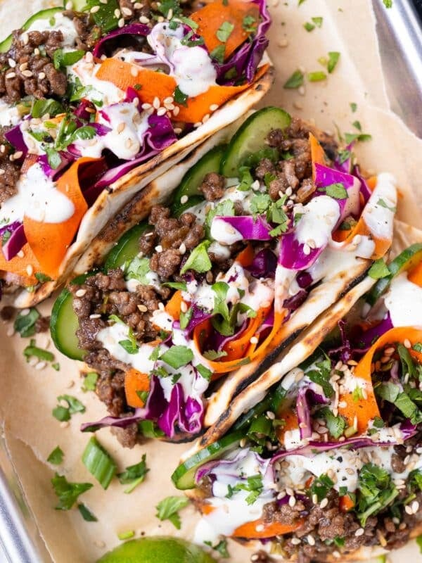 Three Korean Beef Tacos with sesame slaw are on a dinner tray next to lime wedges.