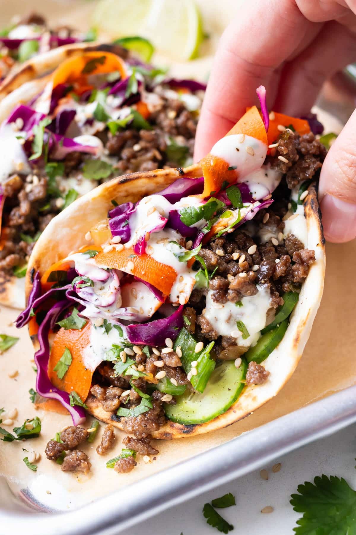 A hand is picking up a Korean Beef Street Taco from a tray with cabbage and carrot slaw and wasabi mayo.