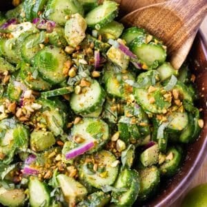 A wooden bowl is full of a cucumber, avocado, and pistachio salad with a ginger-lime dressing.