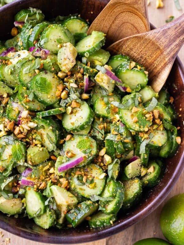 A wooden bowl is full of a cucumber, avocado, and pistachio salad with a ginger-lime dressing.