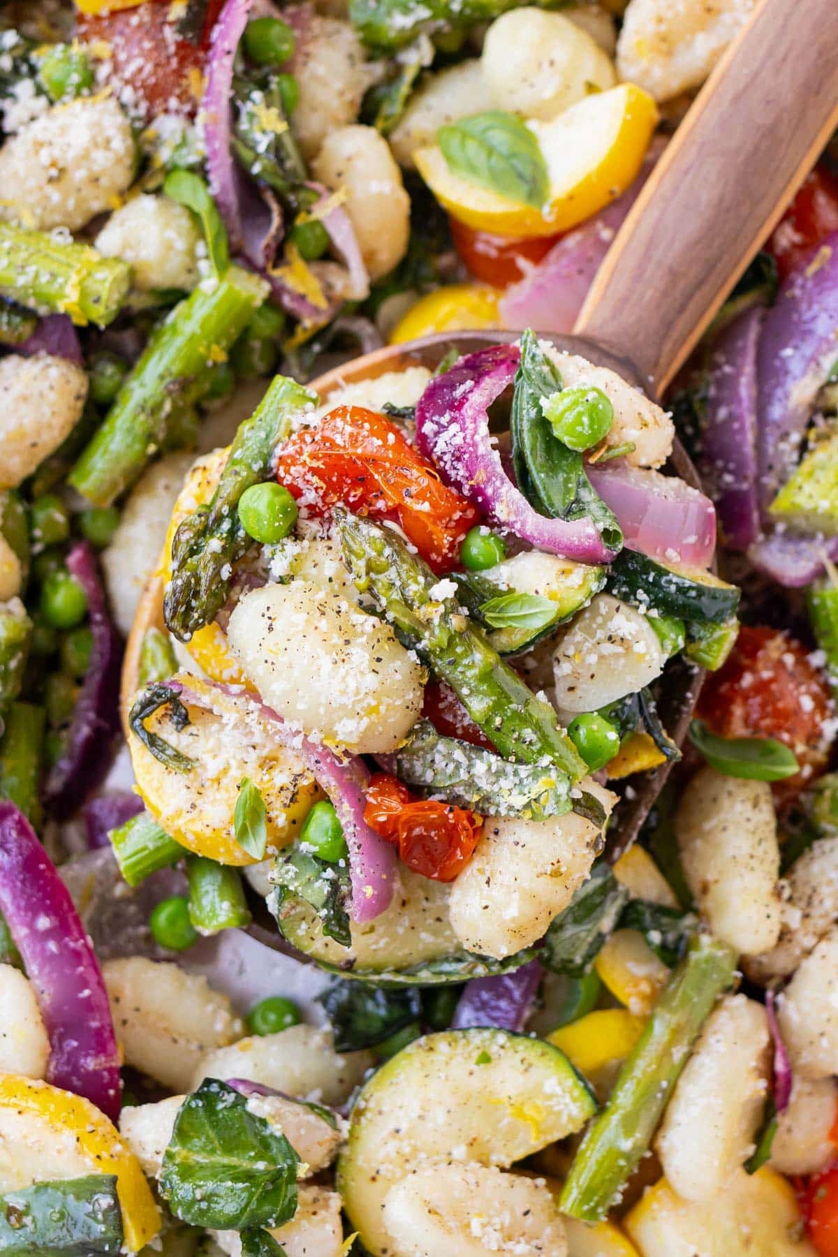 A wooden spoon is scooping up some gnocchi primavera from a sheet pan.
