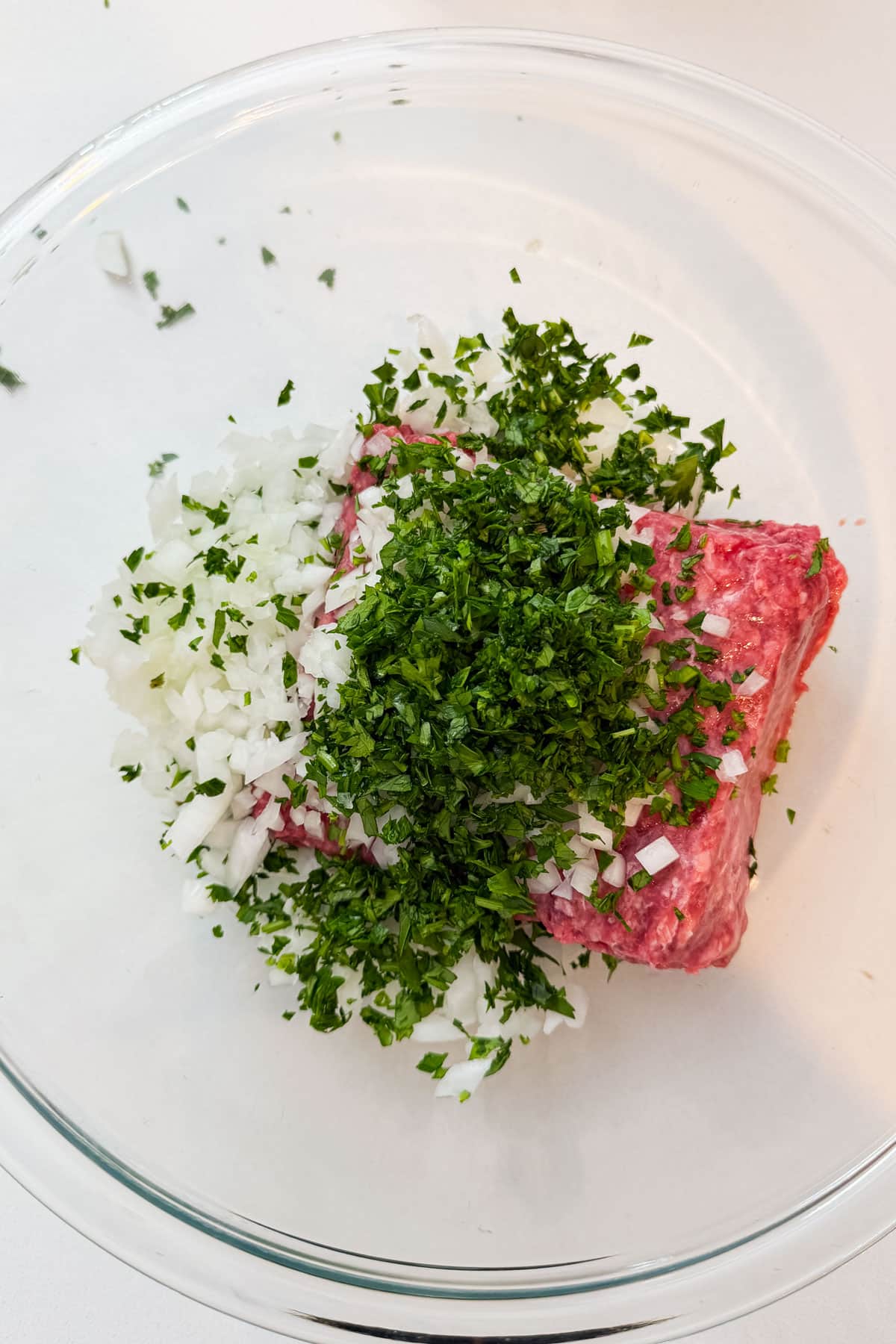 Ground beef topped with diced onion and chopped parsley in a glass mixing bowl for Arayes.