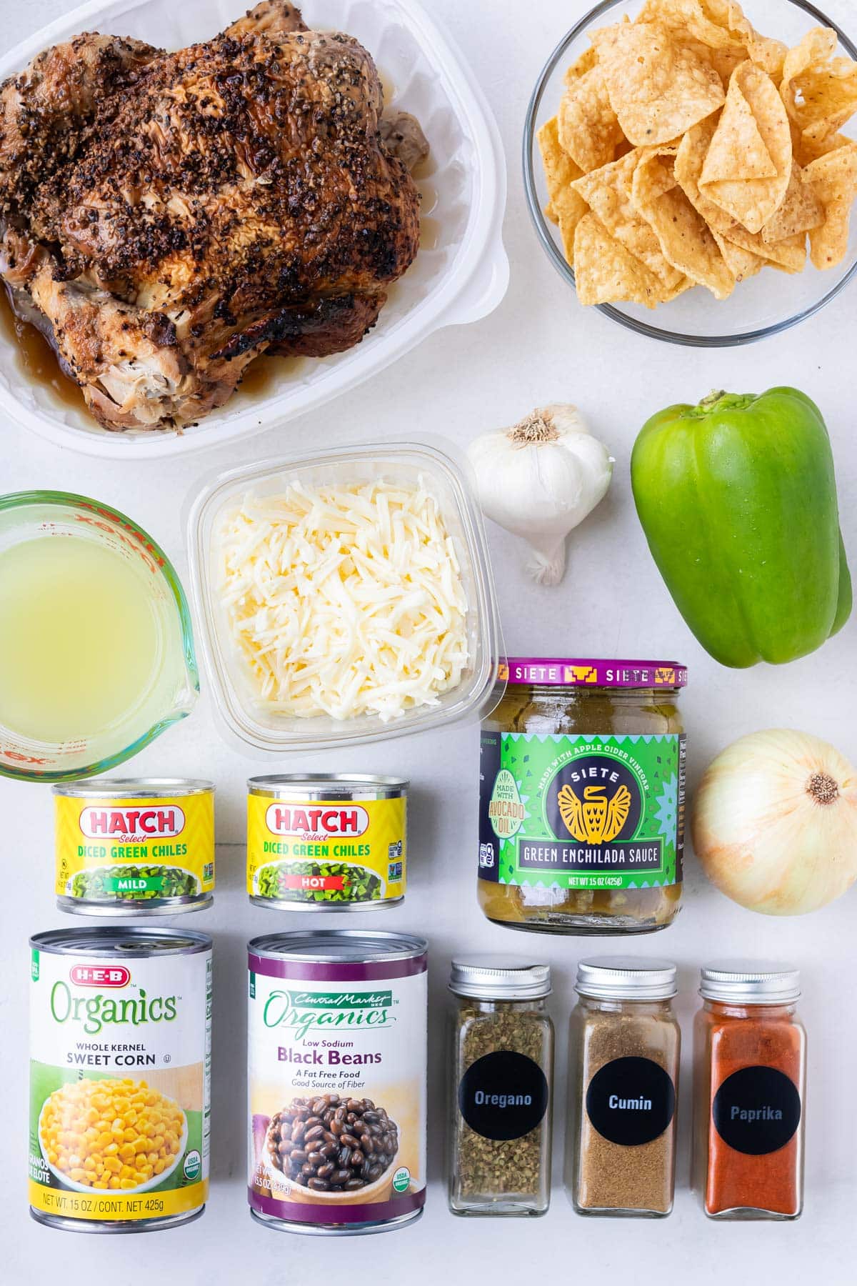 Labeled ingredients for Green Chile Chicken Enchilada Skillet laid out on a white surface: rotisserie chicken, tortilla chips, green bell pepper, garlic, onion, shredded Monterey Jack cheese, chicken broth, green enchilada sauce, corn, black beans, diced green chiles, and spices.