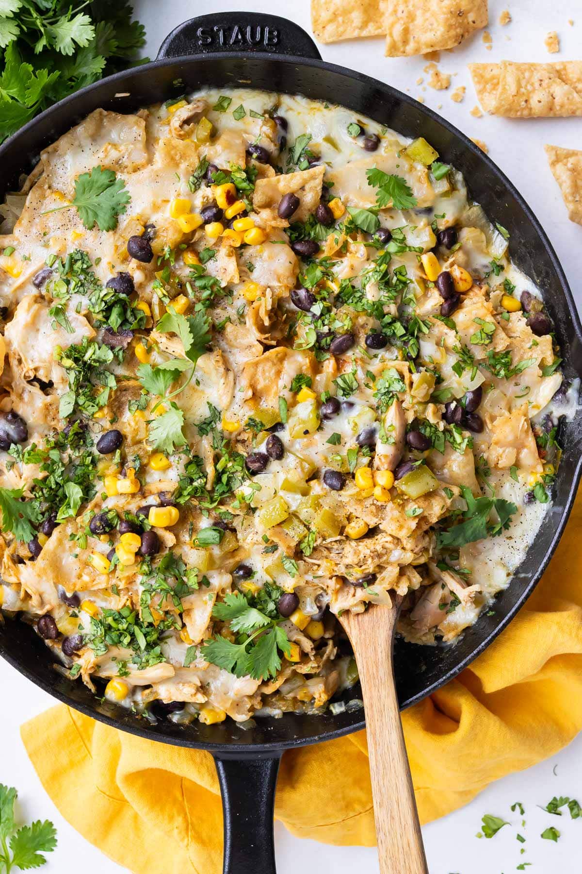 Overhead view of Green Chile Chicken Enchilada Skillet topped with melted Monterey Jack cheese, black beans, corn, and fresh cilantro in a black cast-iron skillet with a wooden spoon and yellow napkin.