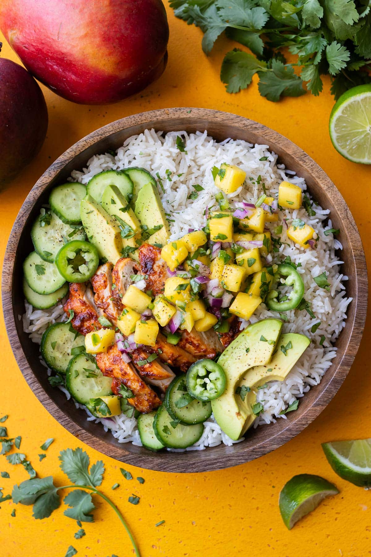 Overhead shot of a wooden bowl filled with Mango Chipotle Chicken Rice Bowls on a yellow background with fresh mango and cilantro.