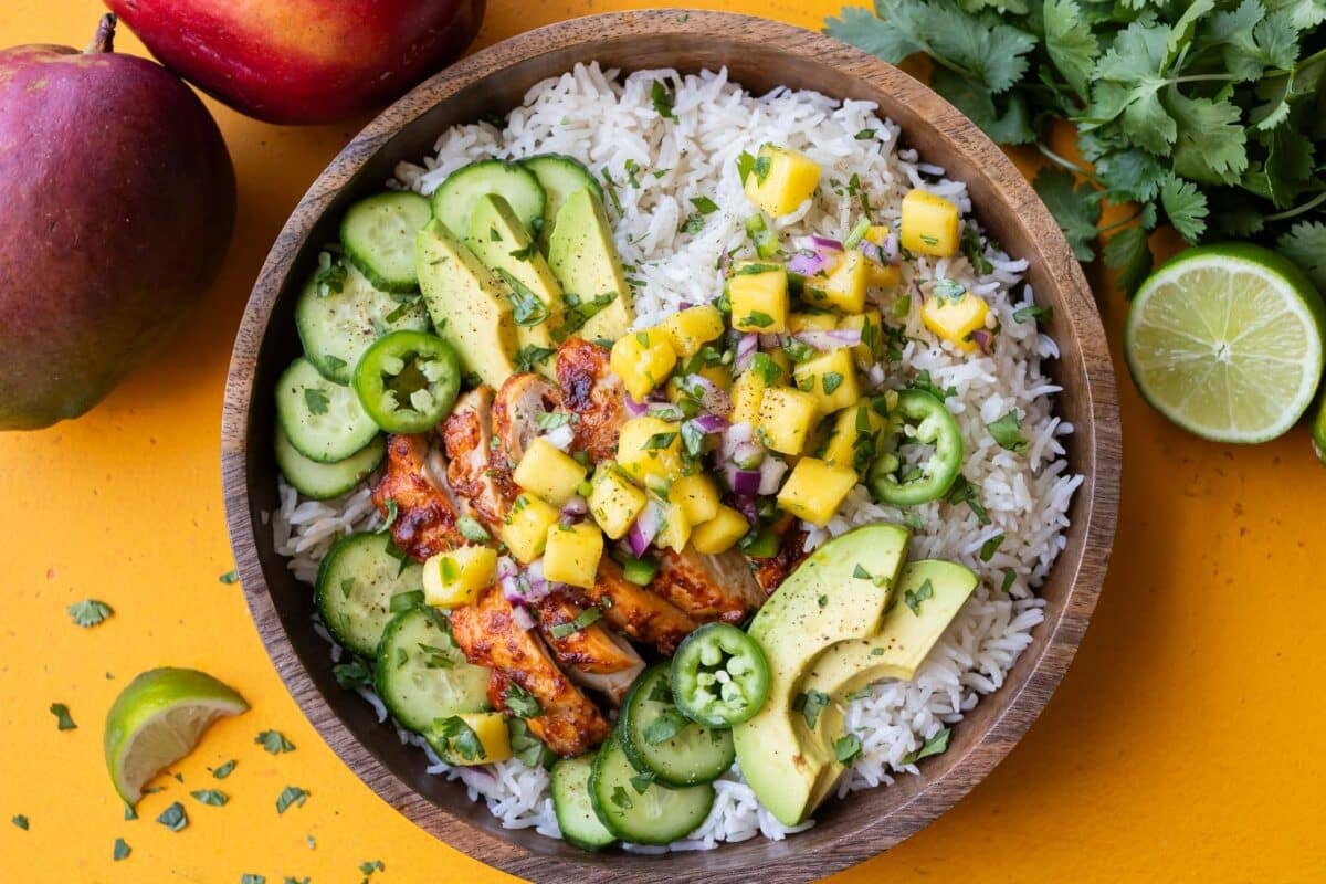A dinner bowl is full of coconut milk rice, chipotle mango chicken thighs, mango salsa, cucumbers and avocado.