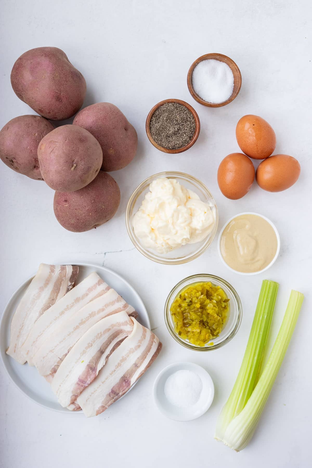 Ingredients for classic potato salad with bacon laid out including red potatoes, eggs, mayonnaise, Dijon mustard, dill pickle relish, celery, and bacon.
