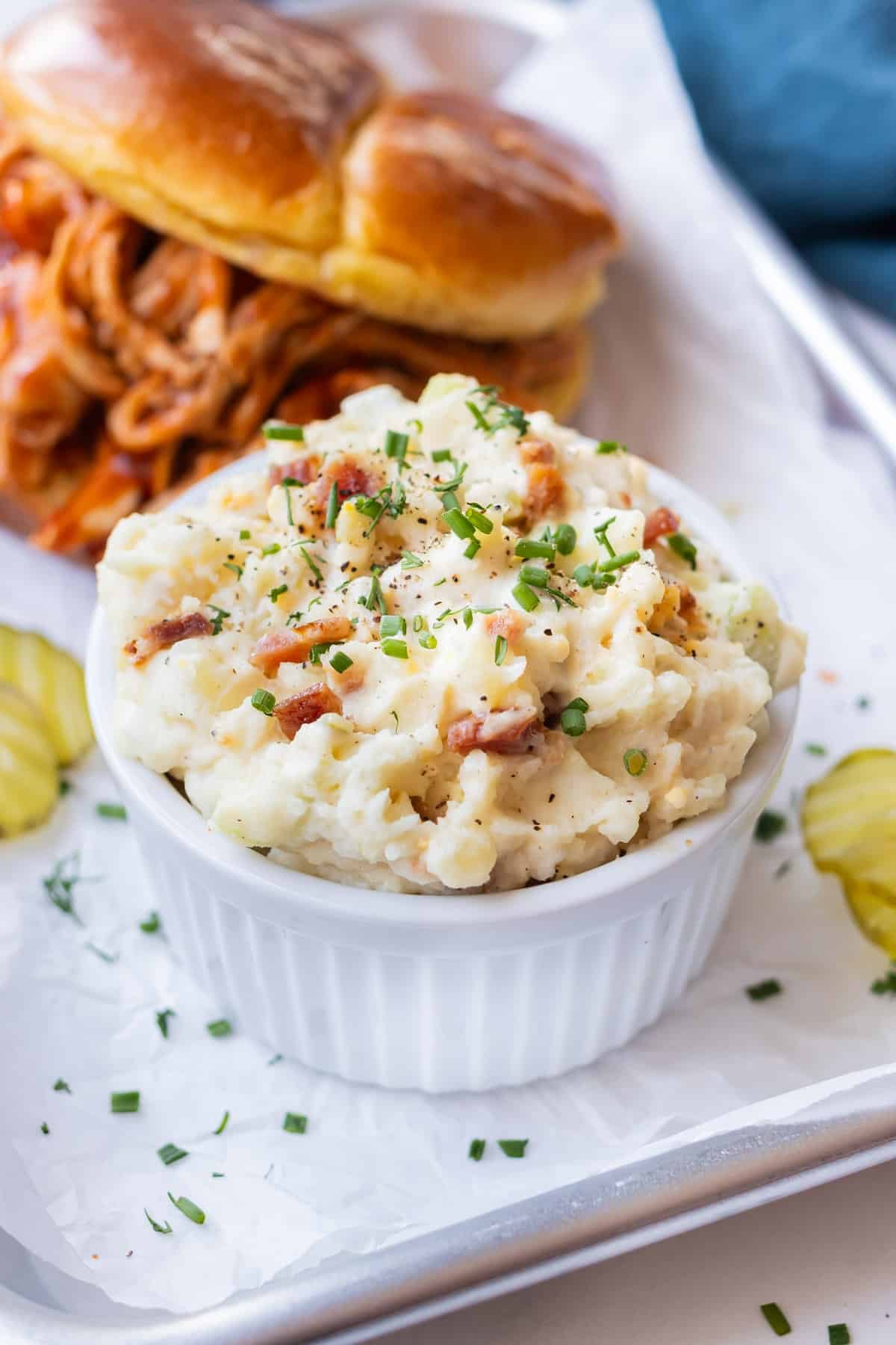 Classic potato salad with bacon served in a white bowl with dill pickles on the side.