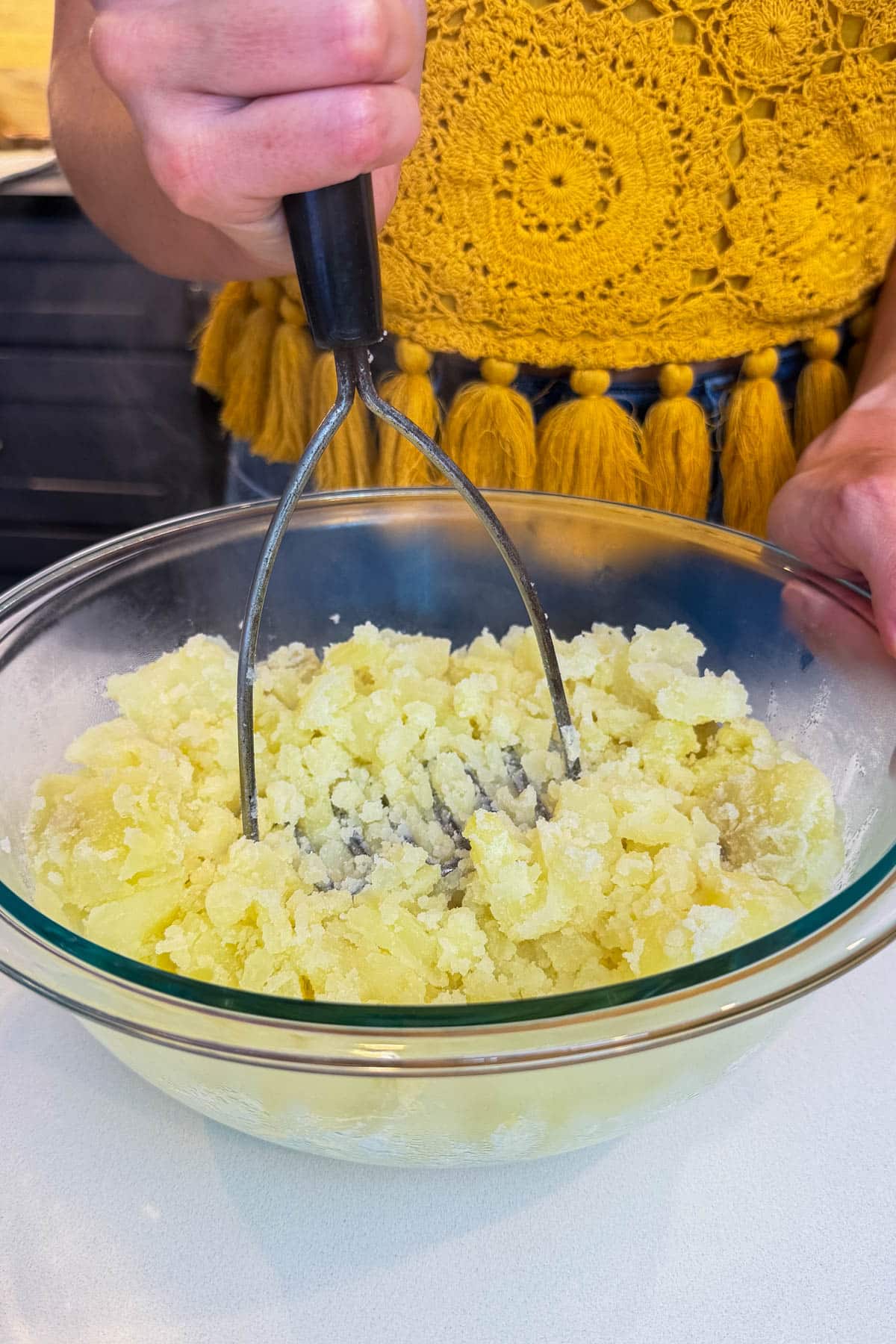 A potato masher gently pressing potatoes in a metal bowl for classic potato salad with bacon.