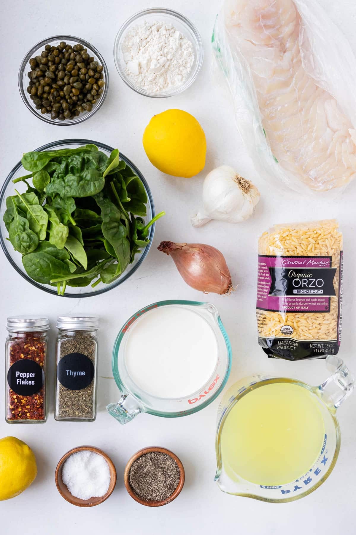 Ingredients for cod piccata with creamy orzo laid out on a white surface including cod fillets, all-purpose flour, capers, lemons, garlic, shallot, orzo pasta, spinach, heavy cream, chicken broth, dried thyme, red pepper flakes, salt, and pepper.