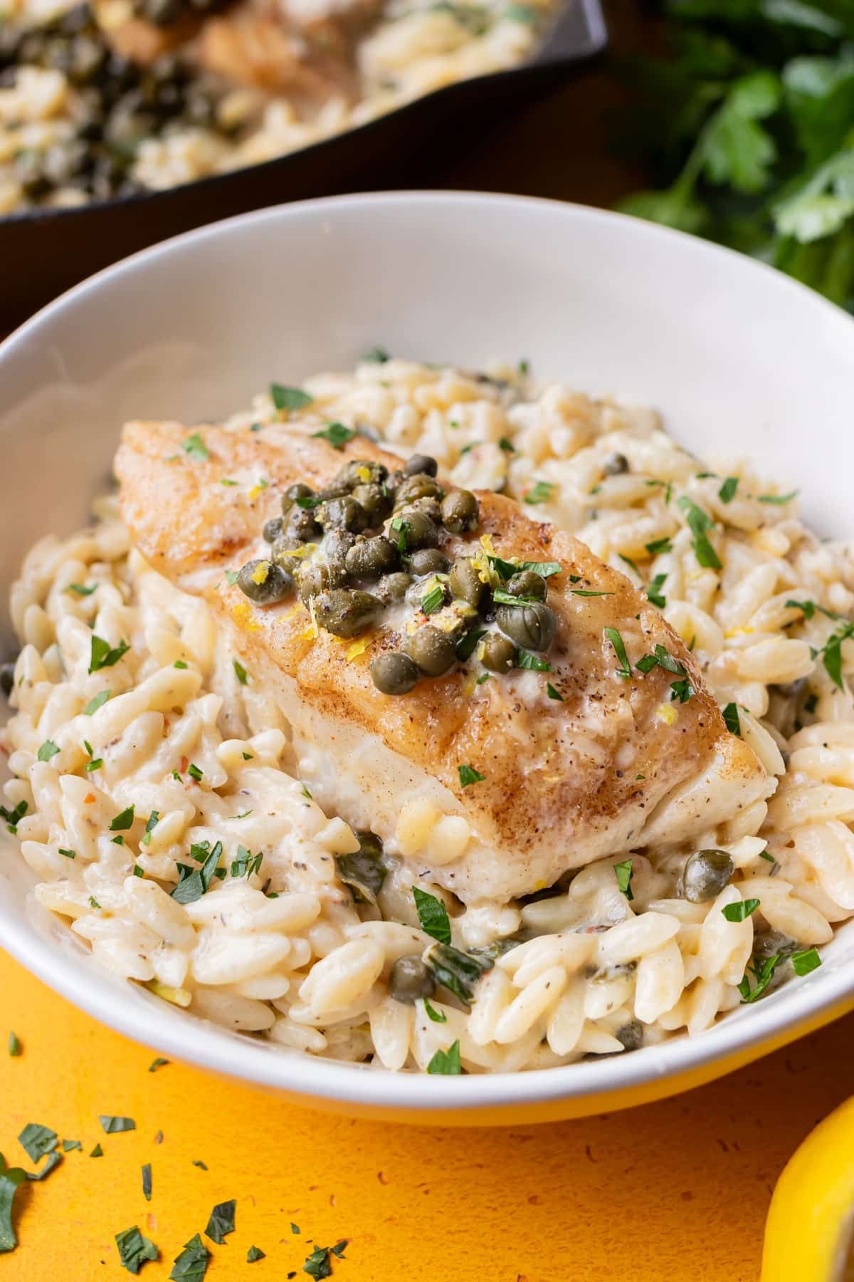 Cod piccata with creamy orzo served in a white bowl, topped with capers and lemon zest.