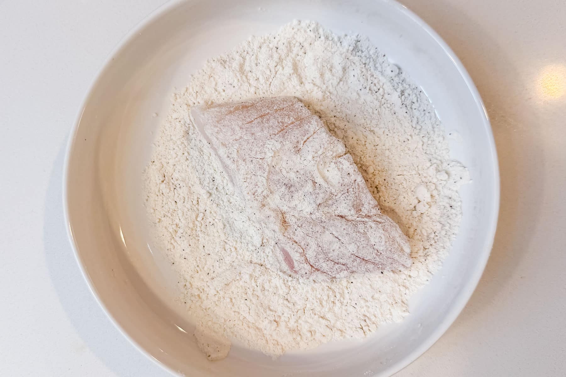 A cod fillet being dredged in flour in a white shallow bowl for cod piccata with creamy orzo.