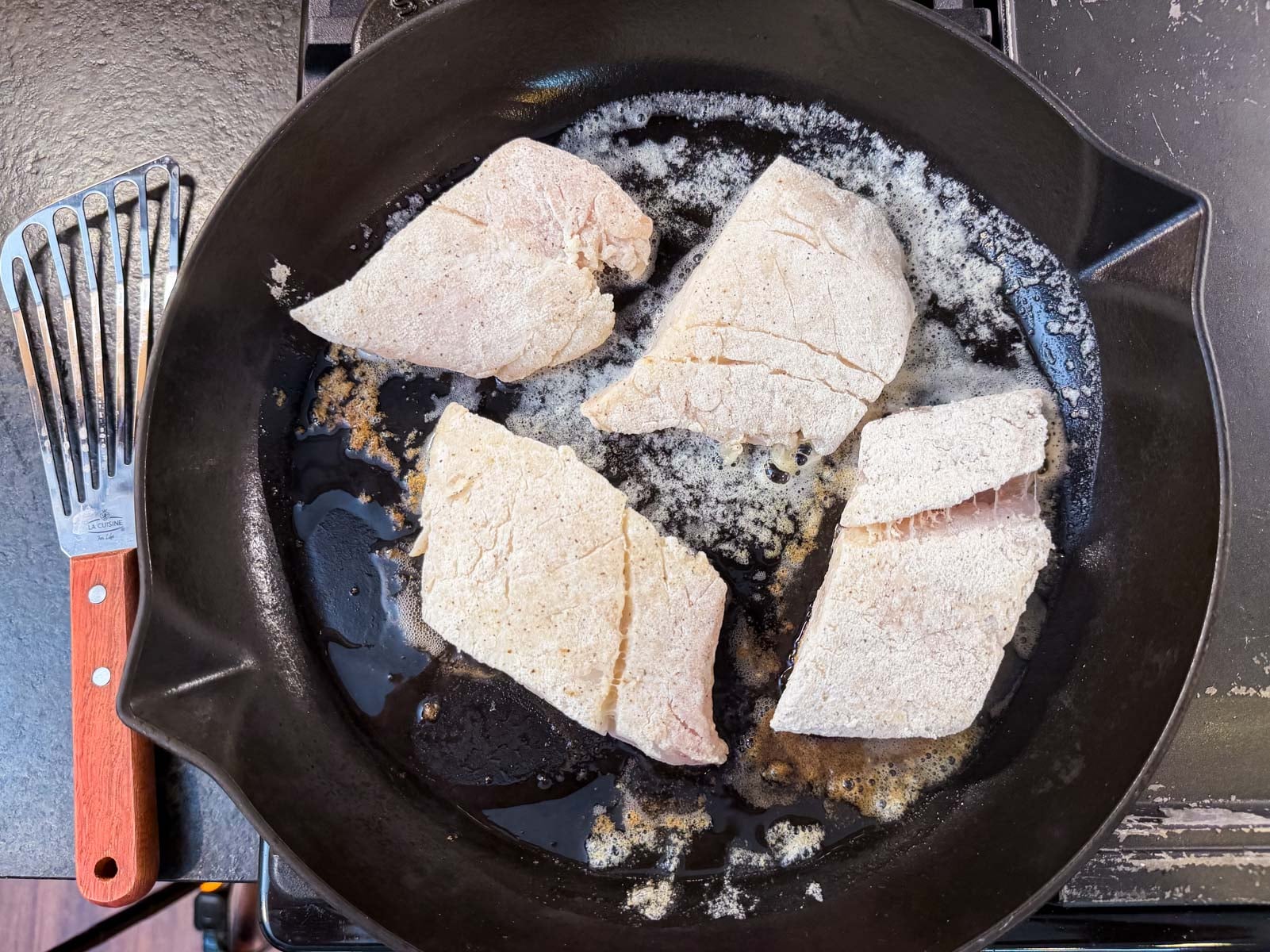 Four dredged cod fillets in a cast iron skillet with melting butter, fish spatula on the side.