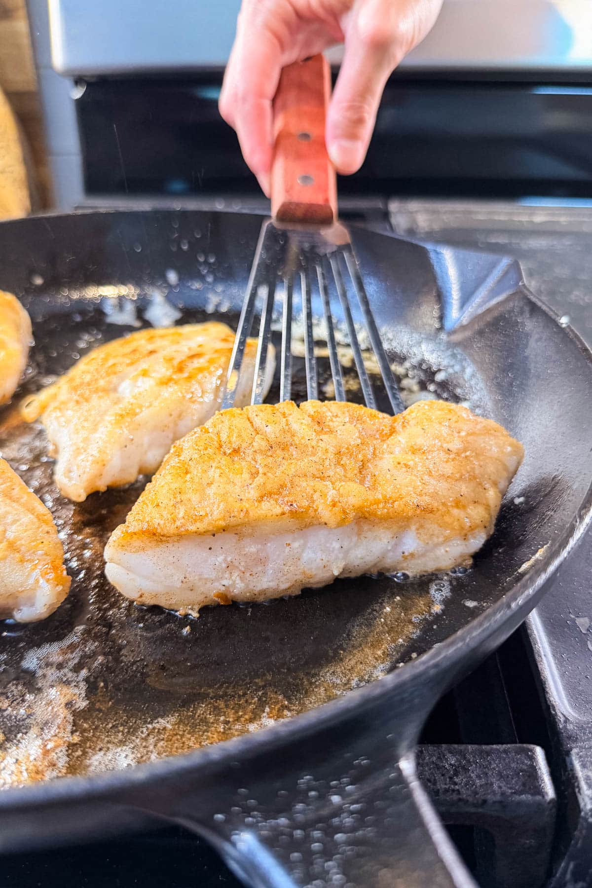 A hand using a fish spatula to flip a golden-brown cod fillet in a cast iron skillet.