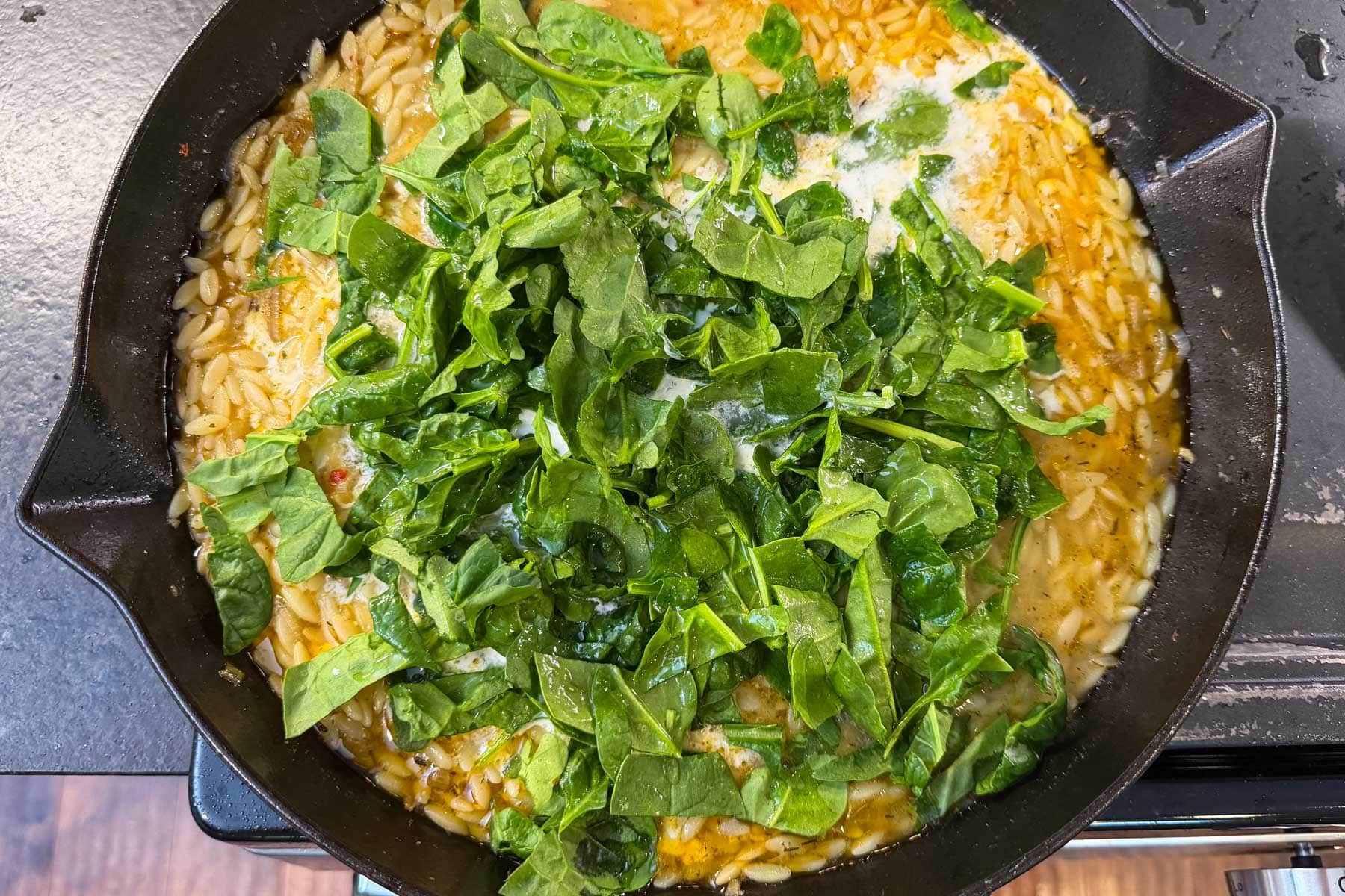 Fresh spinach added on top of creamy orzo in a cast iron skillet, ready to wilt in.
