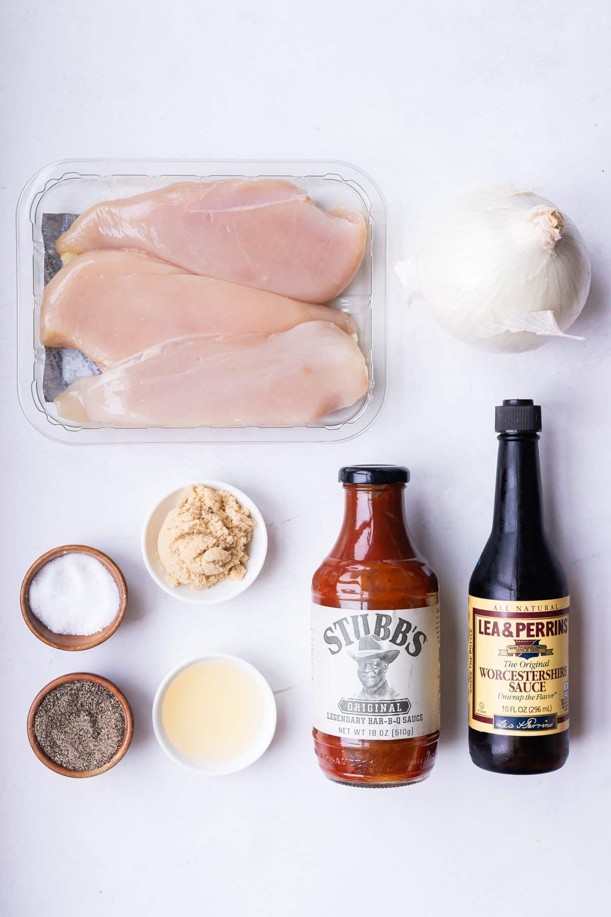 Ingredients for Slow Cooker BBQ Shredded Chicken laid out on a marble surface including chicken breasts, BBQ sauce, brown sugar, onion, garlic, and seasonings.