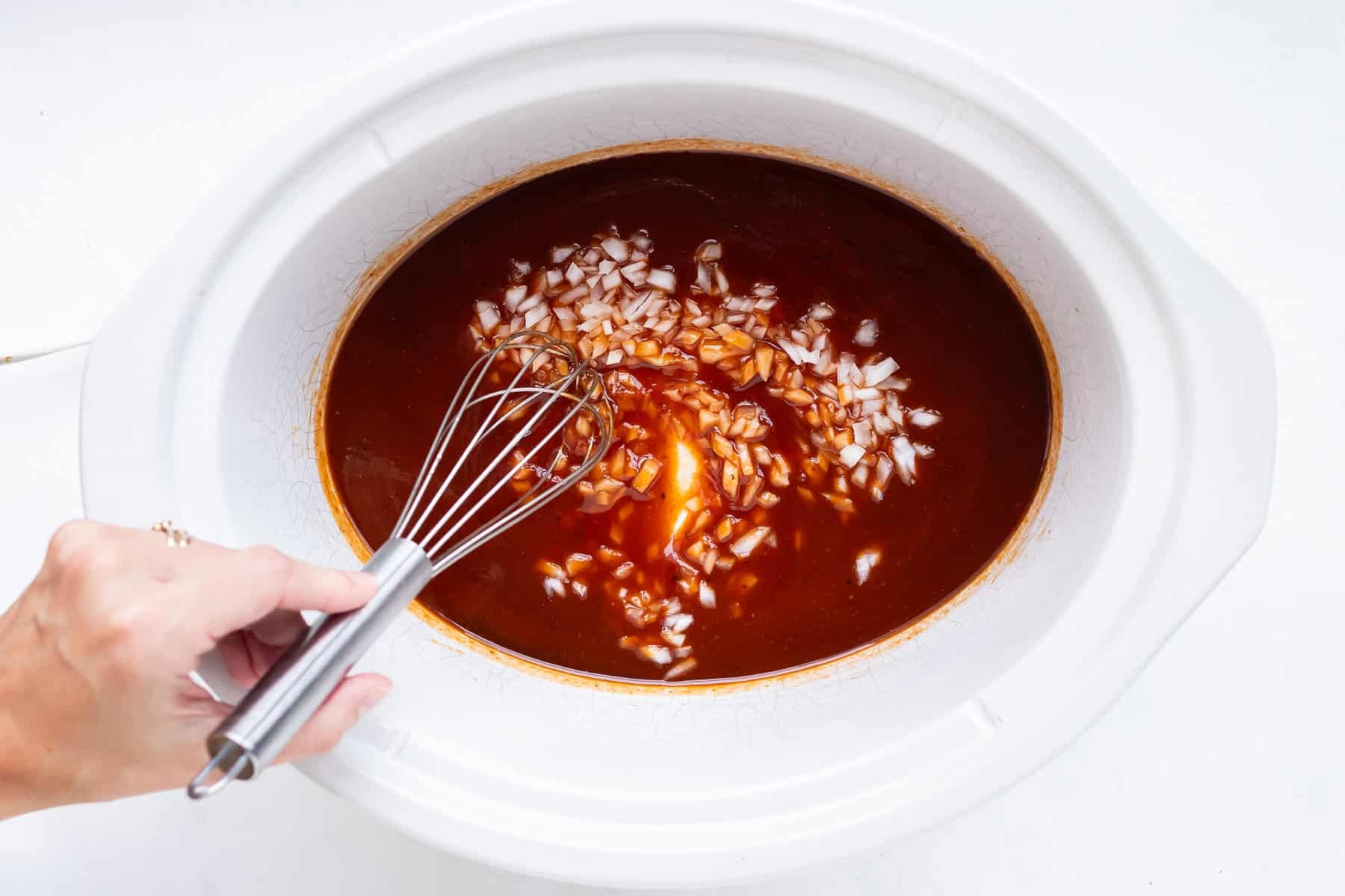 A whisk mixing barbecue sauce, brown sugar, vinegar, and onion together in a slow cooker for Slow Cooker BBQ Shredded Chicken.