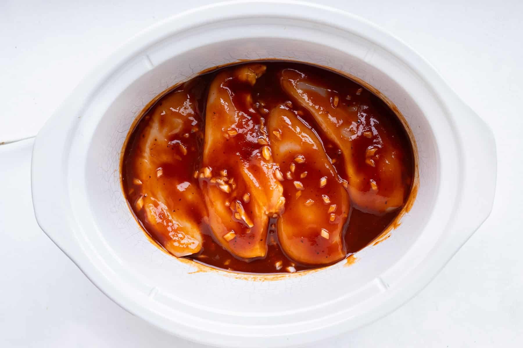 Raw chicken breasts laid in a single layer on top of barbecue sauce in a slow cooker for Slow Cooker BBQ Shredded Chicken.