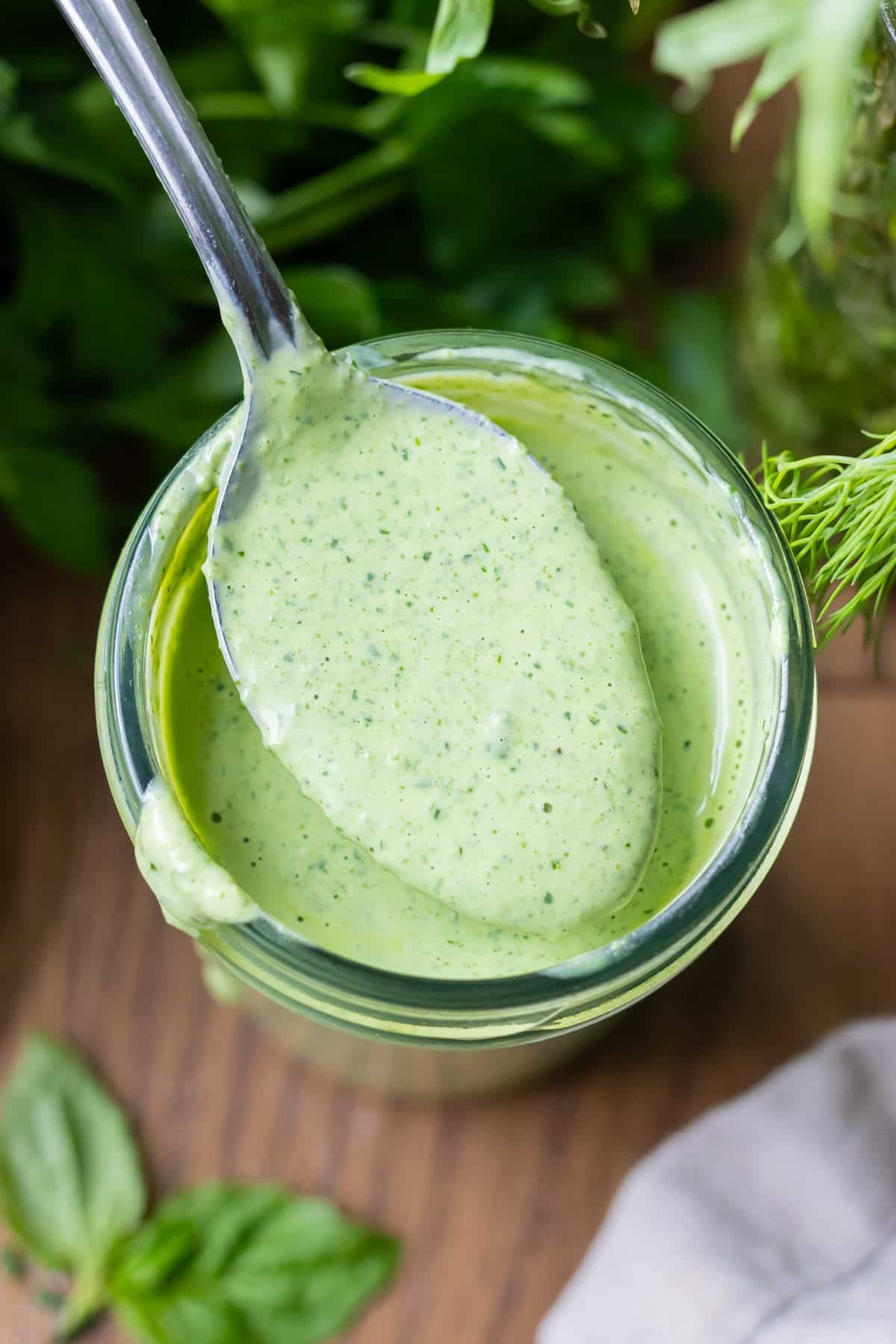 A spoon is held over a jar full of a homemade green goddess dressing recipe with fresh herbs, mayo, and greek yogurt.