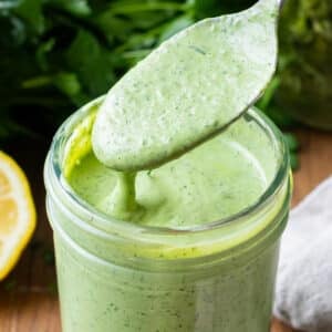 A spoon scoops up a creamy green goddess dressing with fresh herbs from a jar.