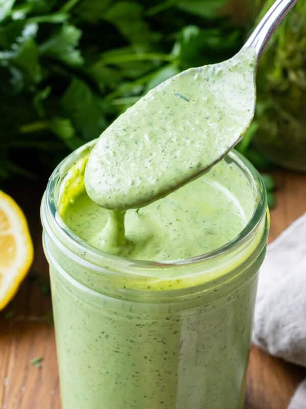 A spoon scoops up a creamy green goddess dressing with fresh herbs from a jar.