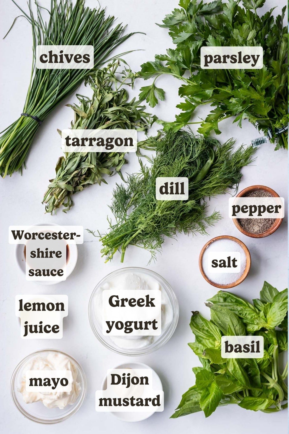 The ingredients for a green goddess salad dressing recipe (chives, basil, parsley, tarragon, dill, yogurt, mayonnaise, worcestershire sauce and dijon mustard) are on a counter.