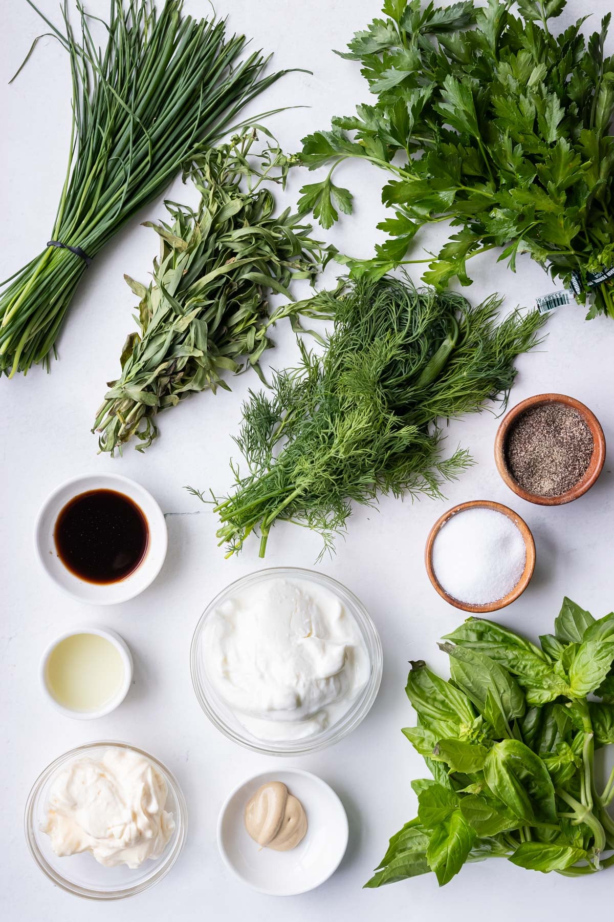 The ingredients for a green goddess salad dressing recipe (chives, basil, parsley, tarragon, dill, yogurt, mayonnaise, worcestershire sauce and dijon mustard) are on a counter.