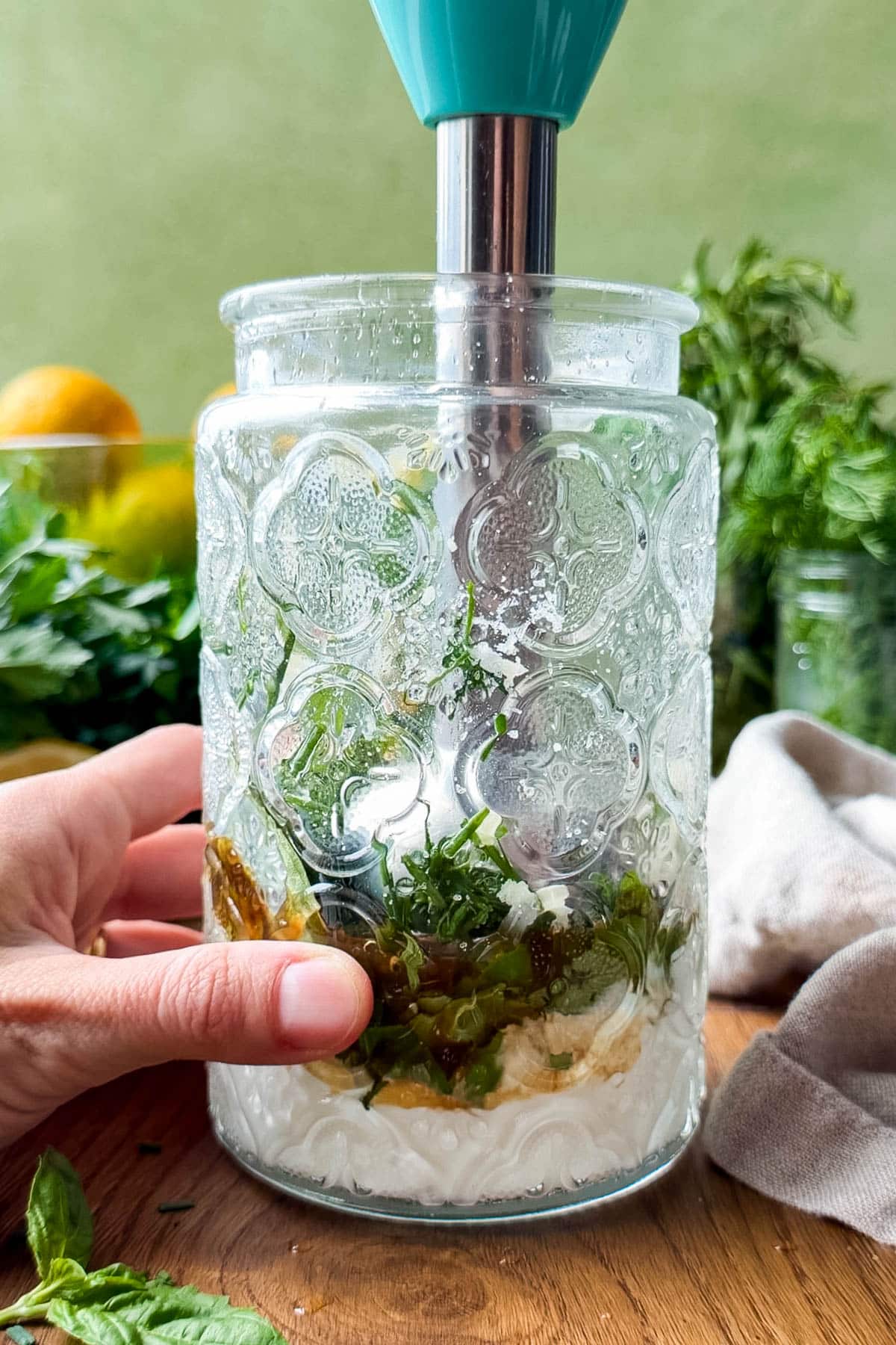 An immersion blender is in a jar full of fresh herbs, yogurt, mayonnaise, and lemon juice blending it into a creamy Green goddess salad dressing recipe.