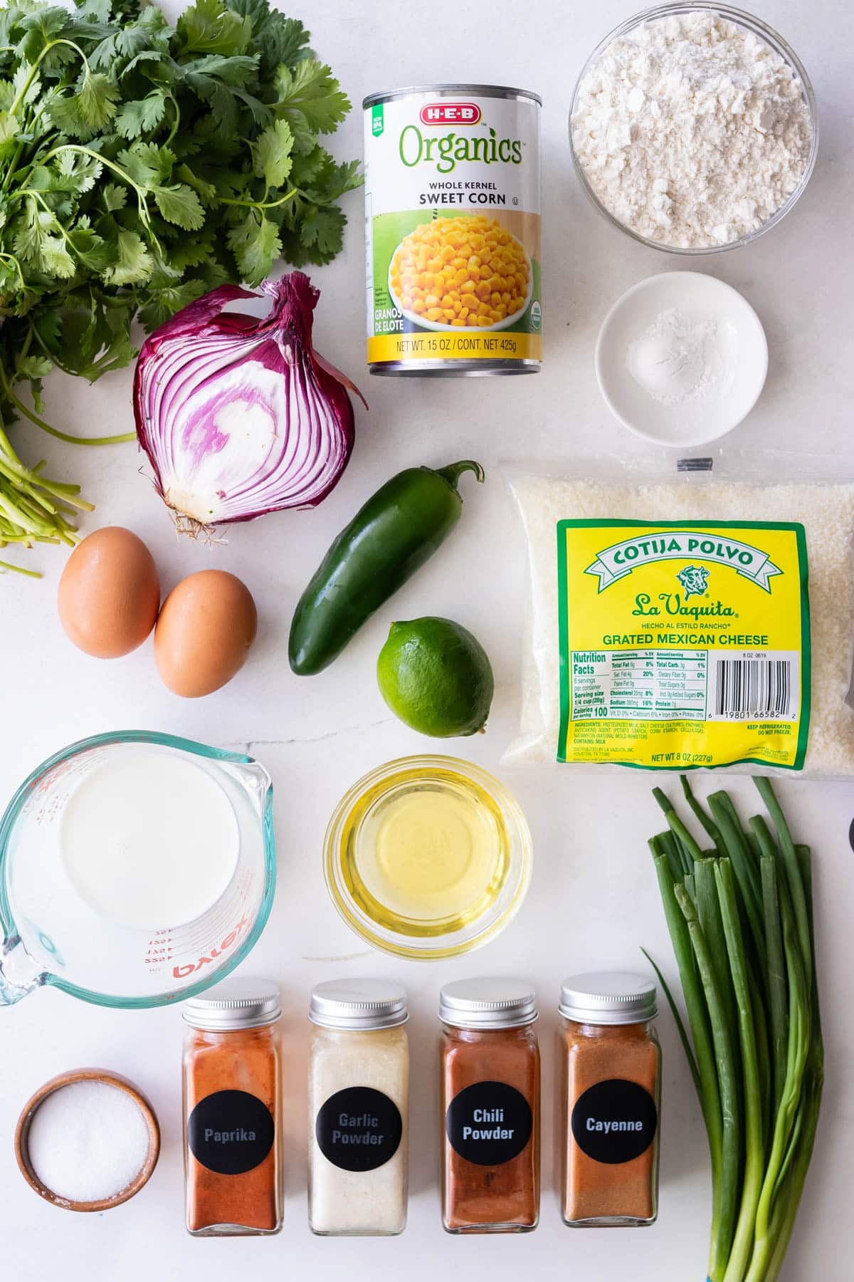 Corn, cilantro, jalapenos, milk, cotija cheese, baking powder, flour, and spices are the ingredients for Mexican street corn fritters on a table.