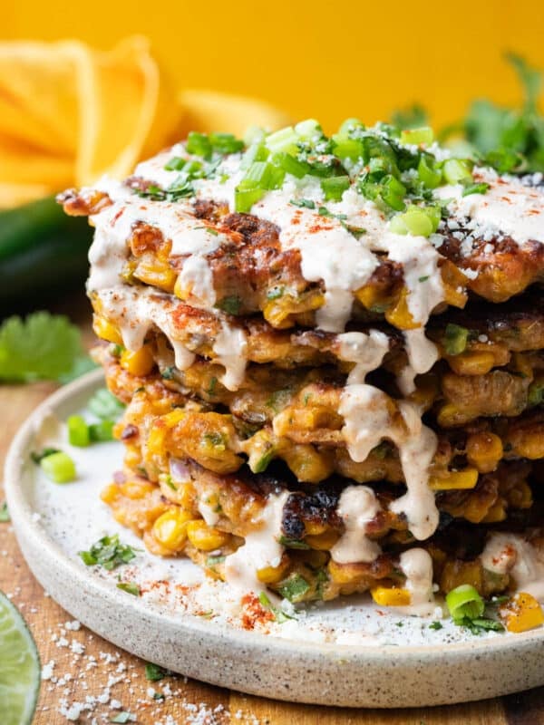 A stack of Mexican street corn fritters are on a plate with a chili lime crema sauce drizzled on top.