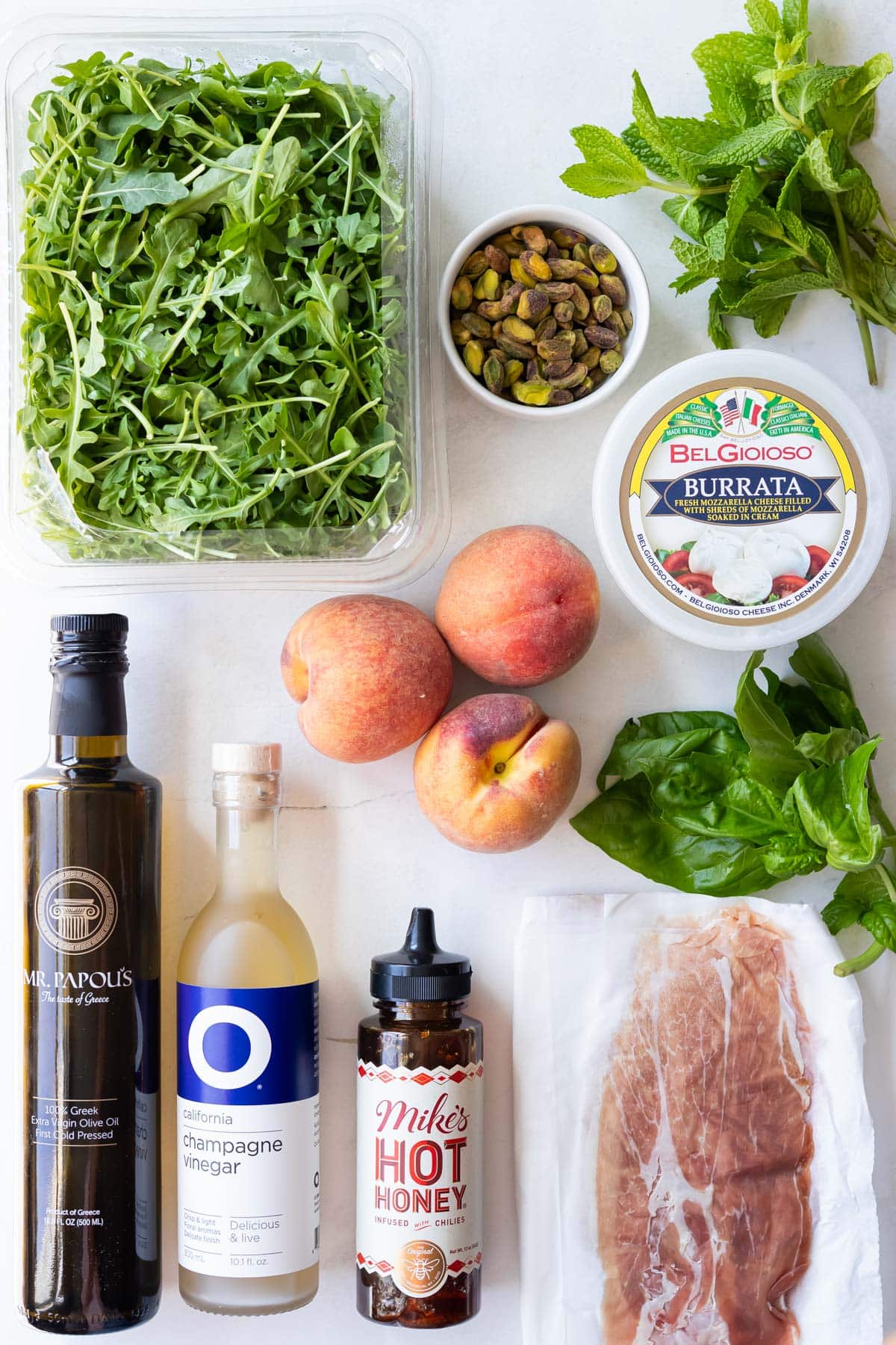 Ingredients for peach burrata salad including arugula, peaches, burrata, prosciutto, pistachios, basil, mint, olive oil, champagne vinegar, and Mike's Hot Honey.