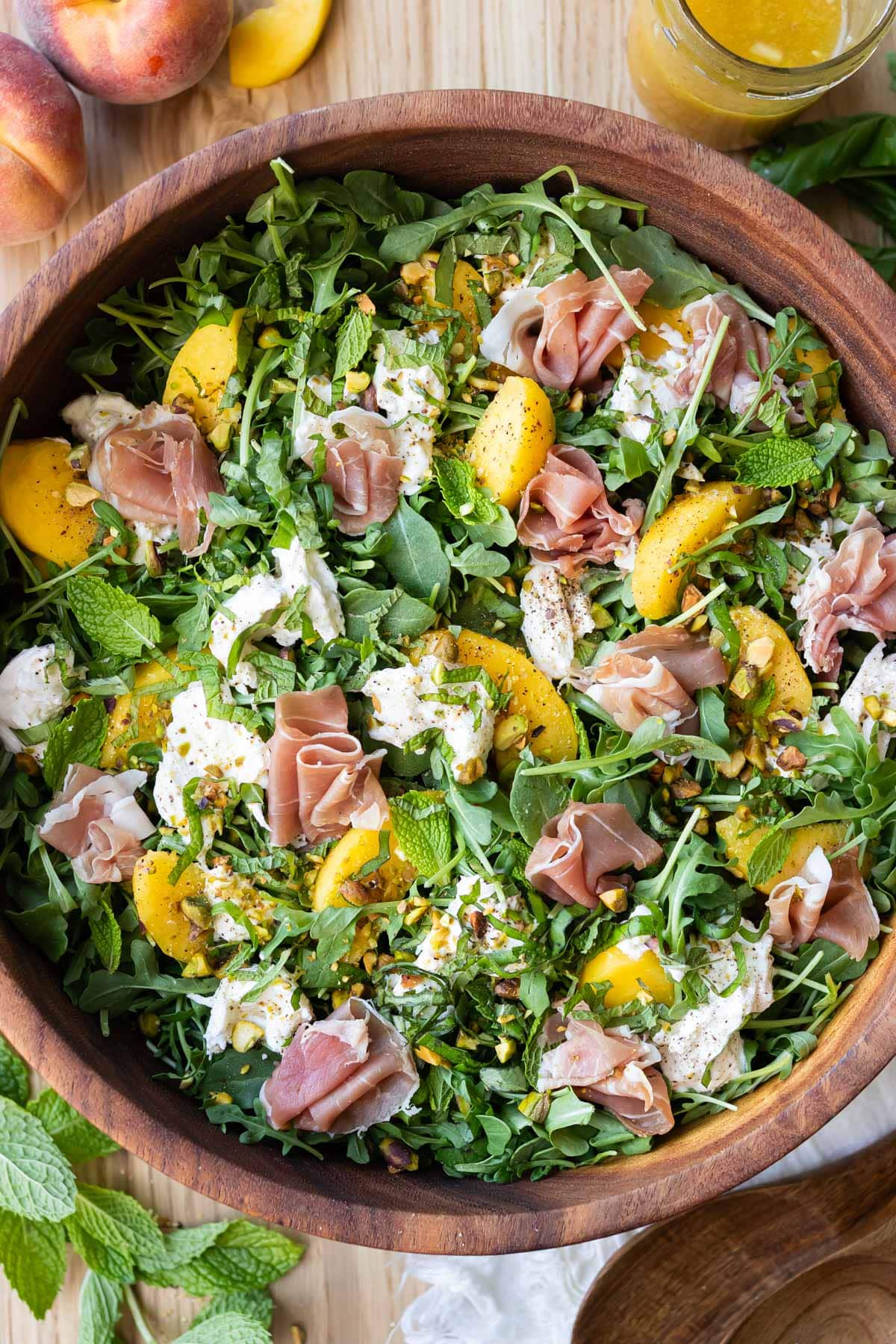 Overhead view of peach burrata salad with arugula, prosciutto, and pistachios in a wooden bowl.