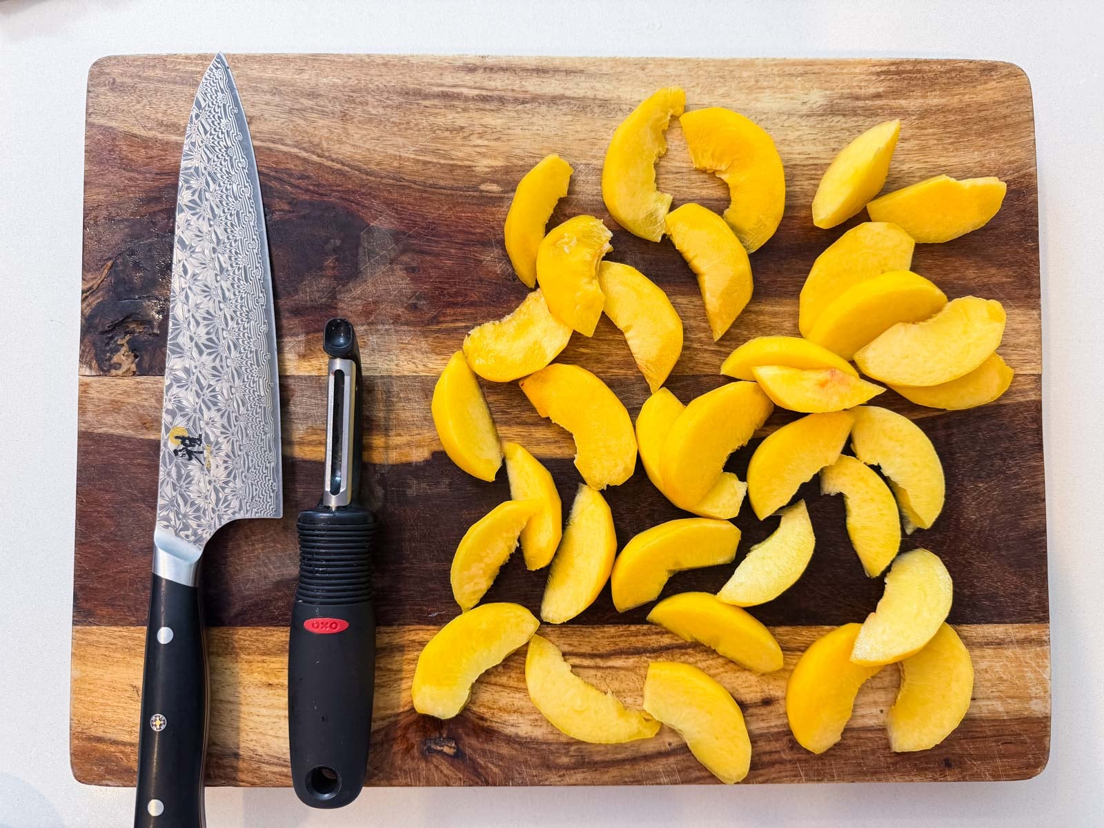 Sliced peach wedges on a wooden cutting board with a chef's knife and serrated vegetable peeler.