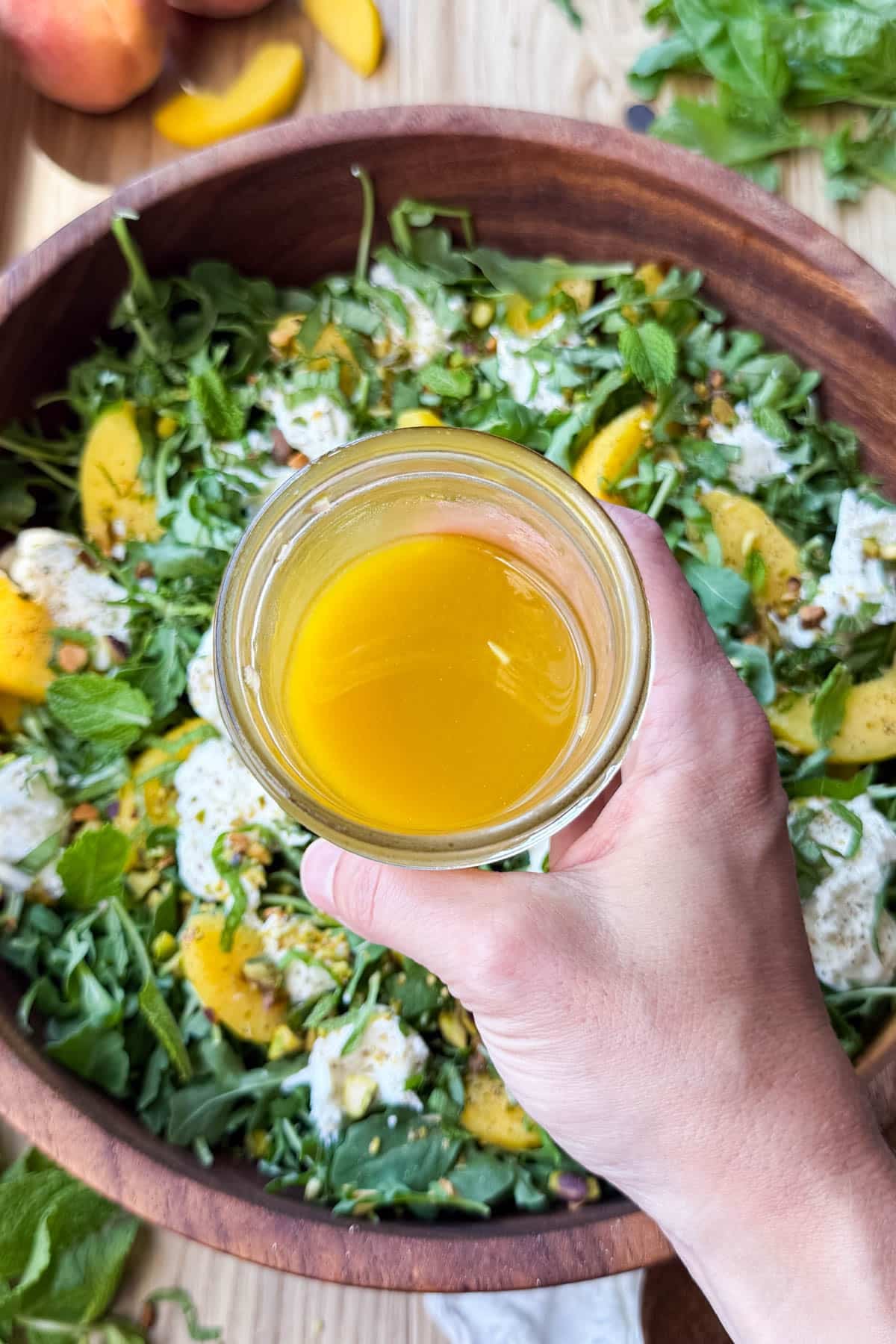 Hand holding a mason jar of hot honey vinaigrette over the assembled peach burrata salad.