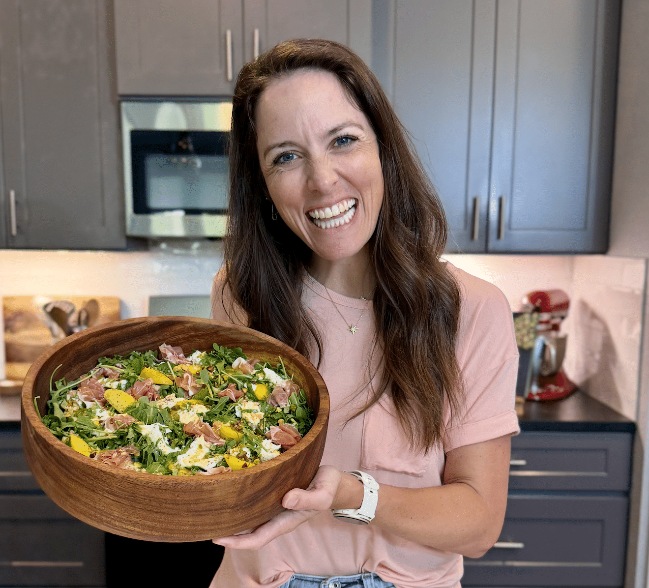 London Lea from Evolving Table is holding a peach burrata salad with arugula in a wooden bowl in her Austin, Texas kitchen.