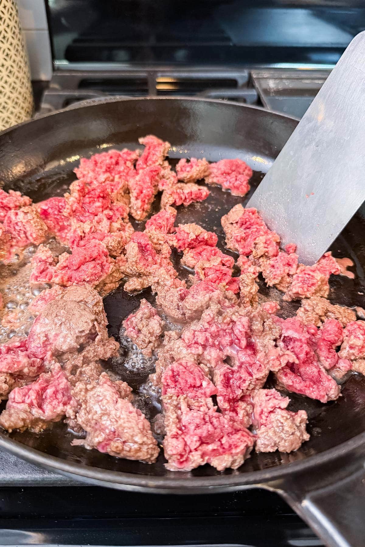 Raw ground beef being browned in a skillet with a metal spatula.