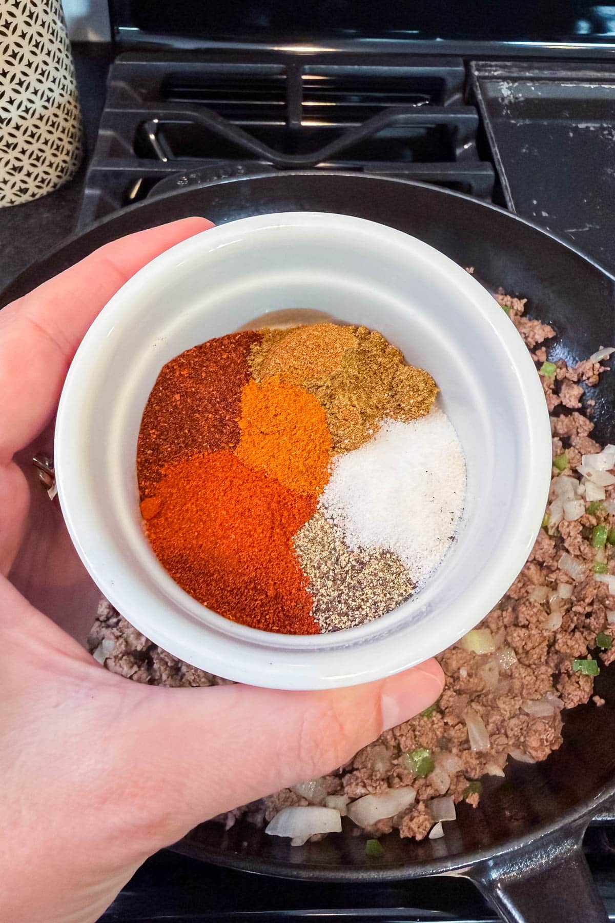 Small bowl of taco spices being added to a skillet of browned ground beef.