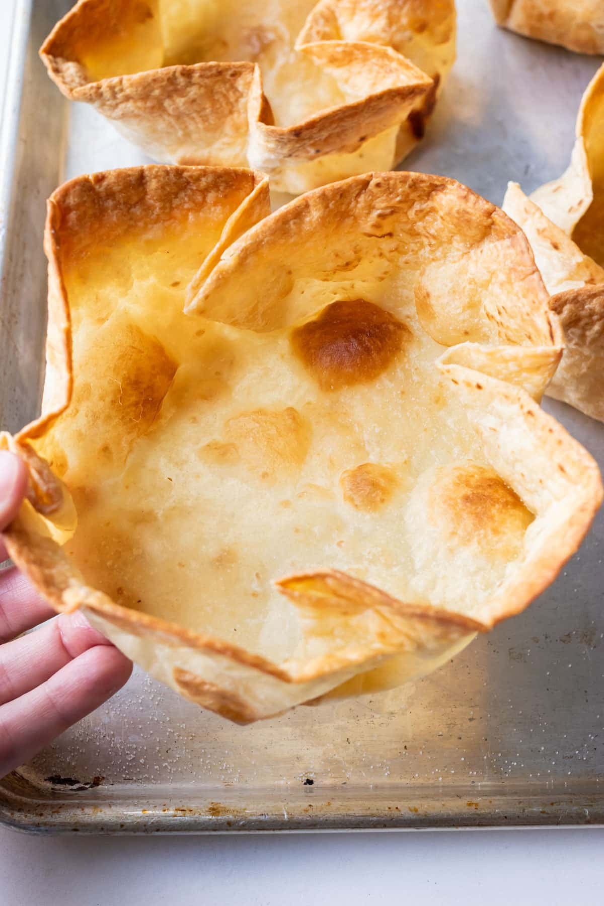 A hand is picking up a bubbly taco salad shell from a baking tray after cooking in the oven.