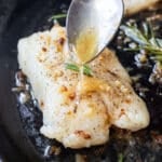 Garlic butter sauce is drizzled over a cod fillet in a cast iron skillet.