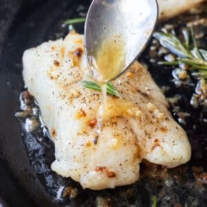 Garlic butter sauce is drizzled over a cod fillet in a cast iron skillet.